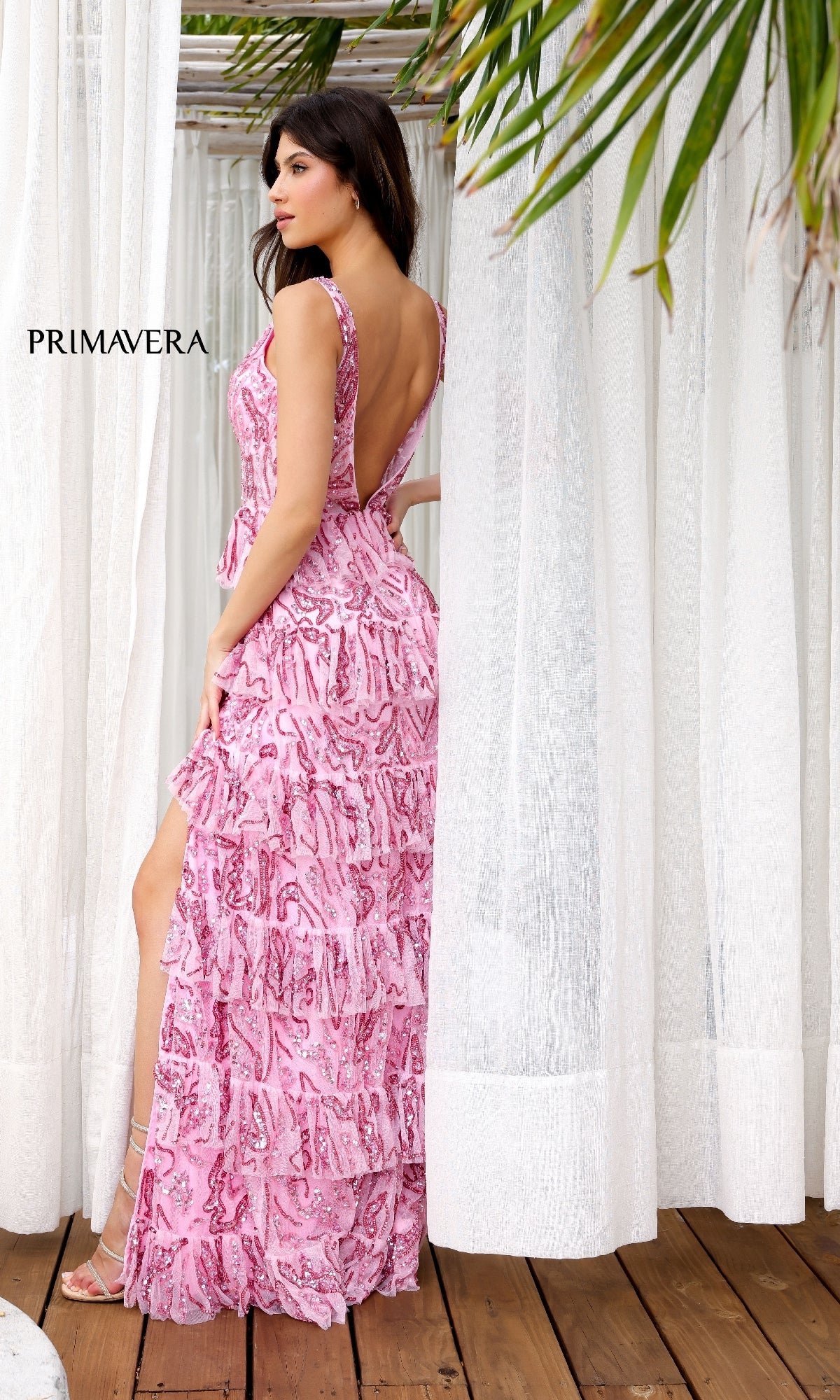 A woman stands on a wooden floor, surrounded by white curtains and tropical leaves, wearing the Primavera 4366 long prom dress—a pink, sleeveless gown with ruffles, a high slit, and tiered layers.
